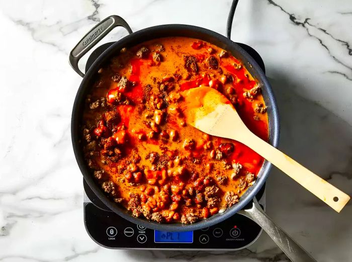 An aerial view showing chili beans, French dressing, and seasoning mix stirred into ground beef in a skillet with a wooden spoon.