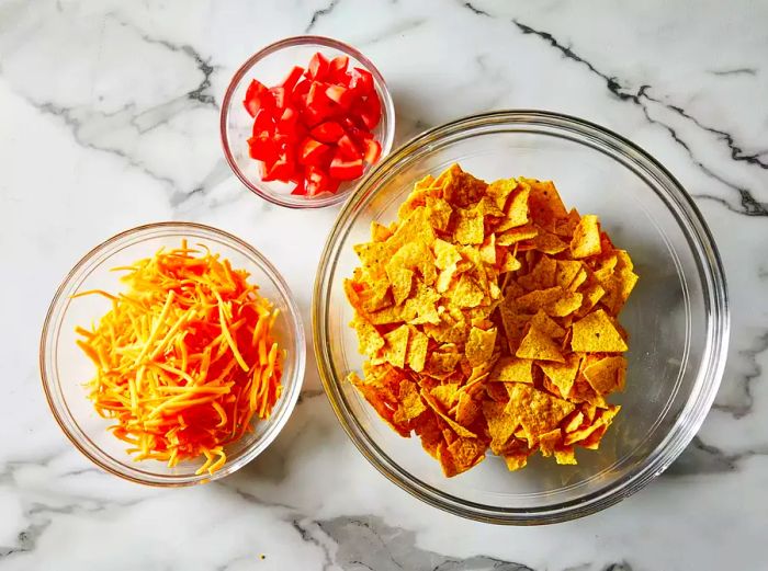 A top-down view of shredded cheese, diced tomatoes, and chips arranged in glass bowls.