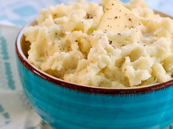 Close-up of a blue-glazed bowl filled with creamy mashed potatoes, topped with freshly cracked pepper and a dollop of butter.