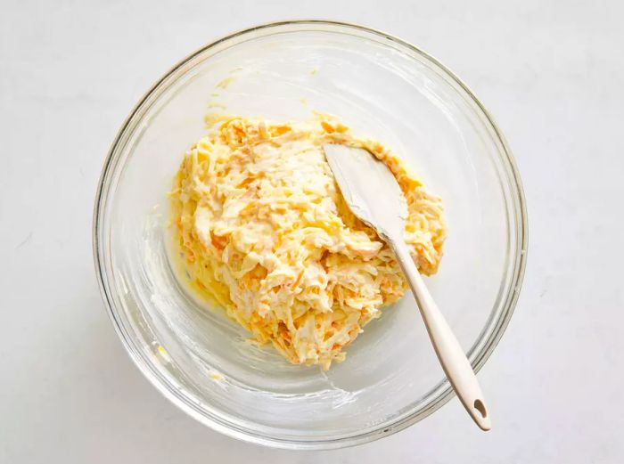 A glass bowl containing a mixture of hash browns, condensed soup, Cheddar cheese, sour cream, onion, and melted butter, stirred with a rubber spatula