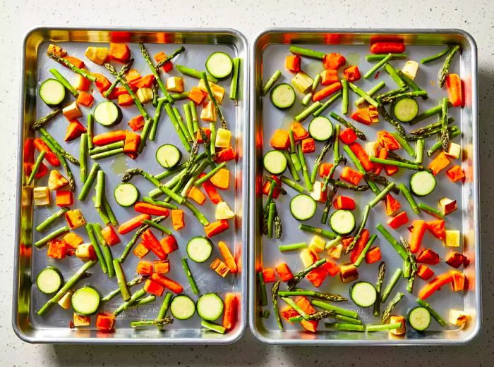 Zucchini and asparagus are added to the partially cooked root vegetables on the two baking sheets.