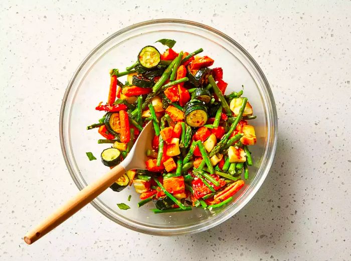 A top-down view of a variety of roasted vegetables being tossed with a peppery mixture.