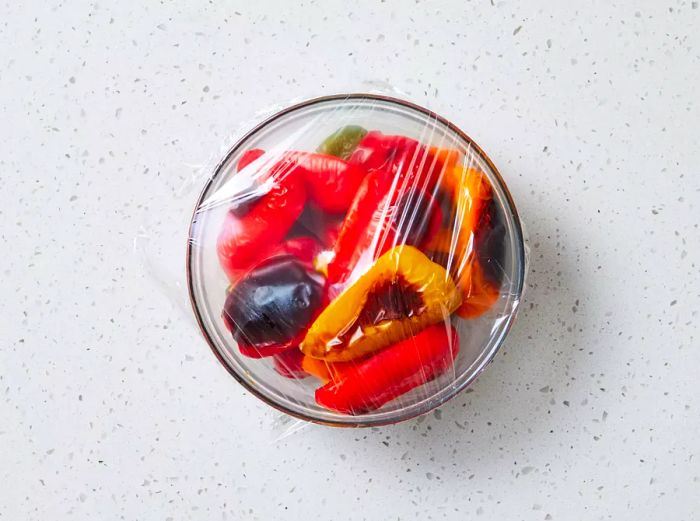 A bowl of quartered, blistered bell peppers covered with plastic wrap