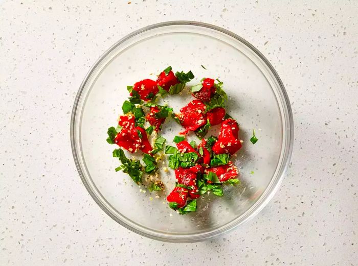 Roasted peppers being mixed with basil, garlic, salt, and pepper in a large bowl.