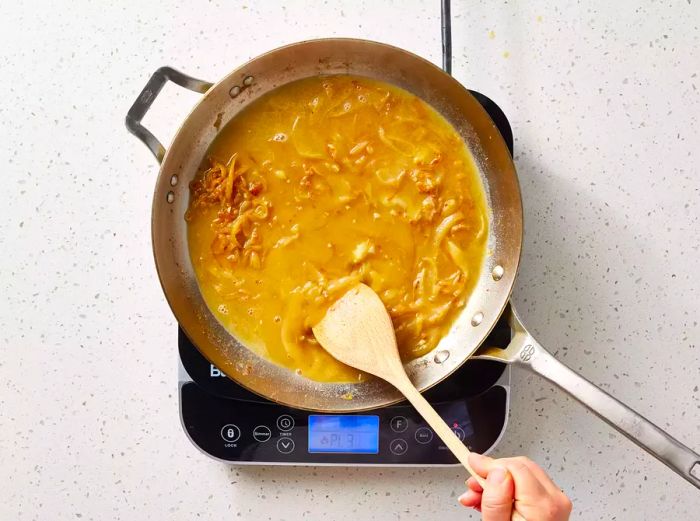 A large pan with a rich brown onion sauce being stirred with a wooden spoon.