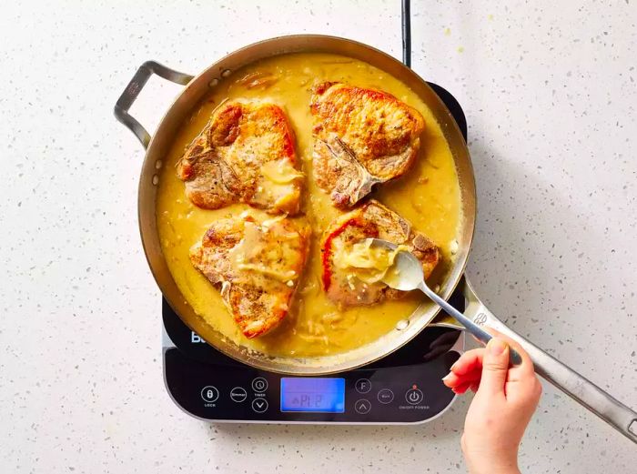 A pan with four pork chops in brown gravy, with a spoon adding caramelized onions on top of each chop.