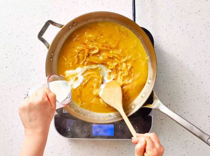A measuring cup with buttermilk being poured into a pan of rich brown onion gravy.