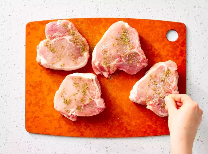 A cutting board featuring four pork chops seasoned with poultry seasoning, salt, and pepper.