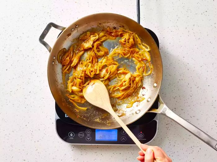 A large skillet with caramelized onions being stirred with a wooden spoon.
