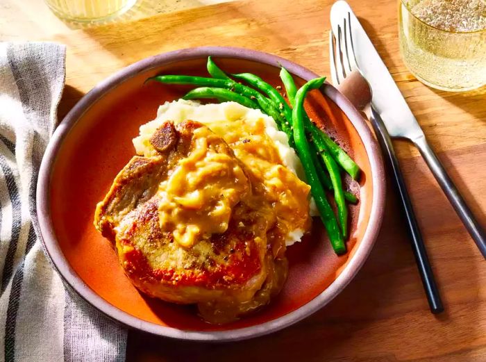 A plate featuring a pork chop smothered in savory sauce, paired with mashed potatoes and green beans.