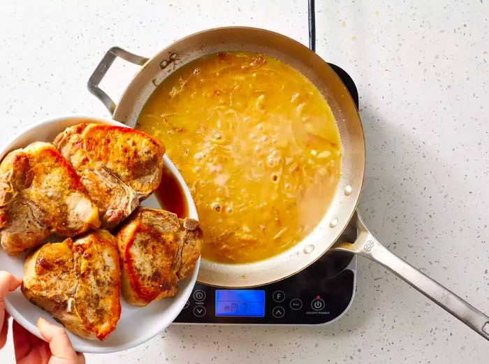 The liquid from a plate of browned pork chops being poured into a pan with brown onion sauce.