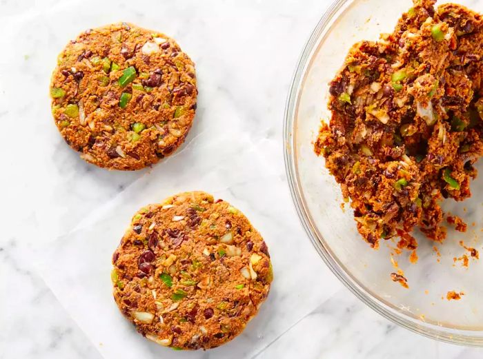 Two black bean veggie burger patties next to a clear bowl containing more burger mixture.