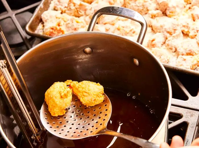 A large pot filled with hot oil, with golden-brown chicken nuggets being lifted out using a metal skimmer.