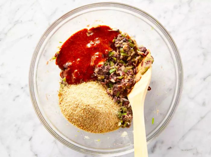 The black bean veggie burger mixture, breadcrumbs, and chili spice blend in a clear bowl being stirred with a wooden spatula.