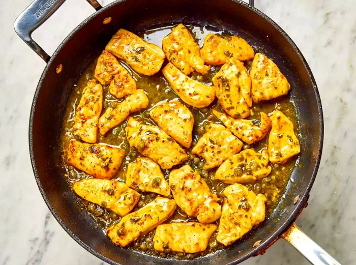A skillet with pieces of browned chicken breasts simmering in a white wine sauce with capers.