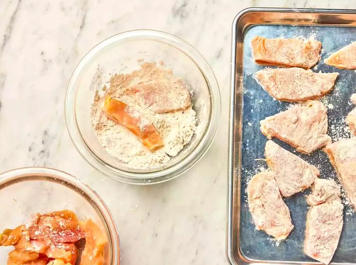 A bowl filled with seasoned flour and small pieces of chicken being coated, alongside a baking sheet with dredged chicken breast pieces.