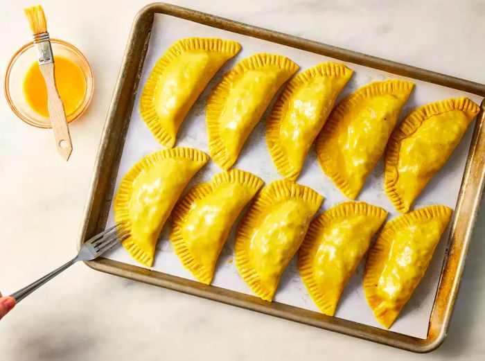 A baking sheet lined with parchment paper, featuring egg-washed Jamaican beef patties sealed with a fork.