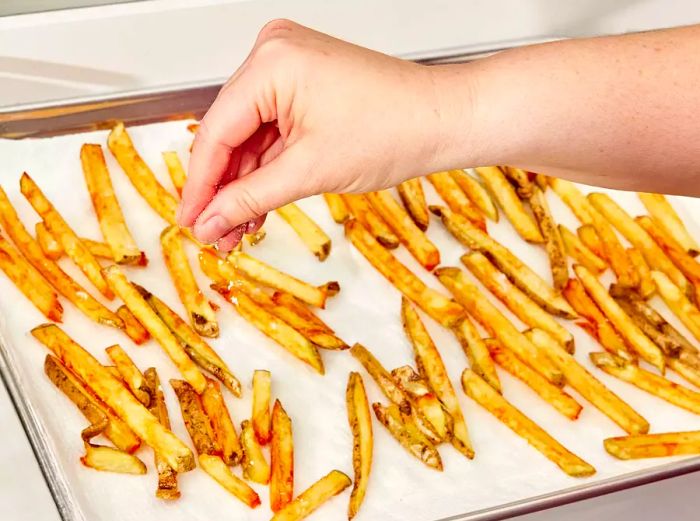 A high-angle shot of fries on a baking sheet being sprinkled with salt