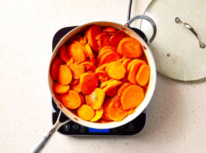 A large pot filled with sliced, candied sweet potatoes next to a glass lid.