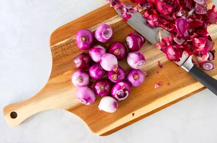 Freshly peeled whole shallots on a wooden cutting board, ready to be used, with a chef's knife nearby.
