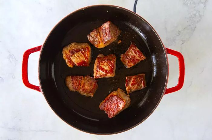Seared cubes of beef cooking in an enameled cast iron skillet, with a golden-brown crust forming.