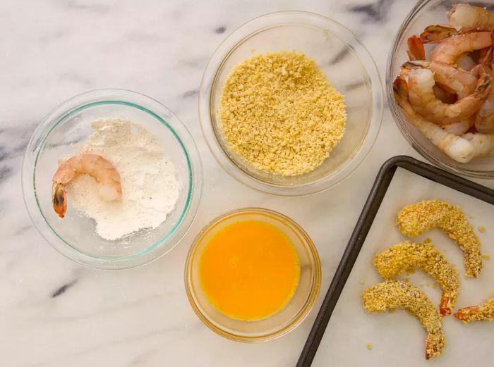 A raw shrimp placed in a small bowl of flour, next to a bowl of beaten eggs, a bowl of breadcrumbs, and a baking sheet lined with parchment paper, holding the breaded shrimp