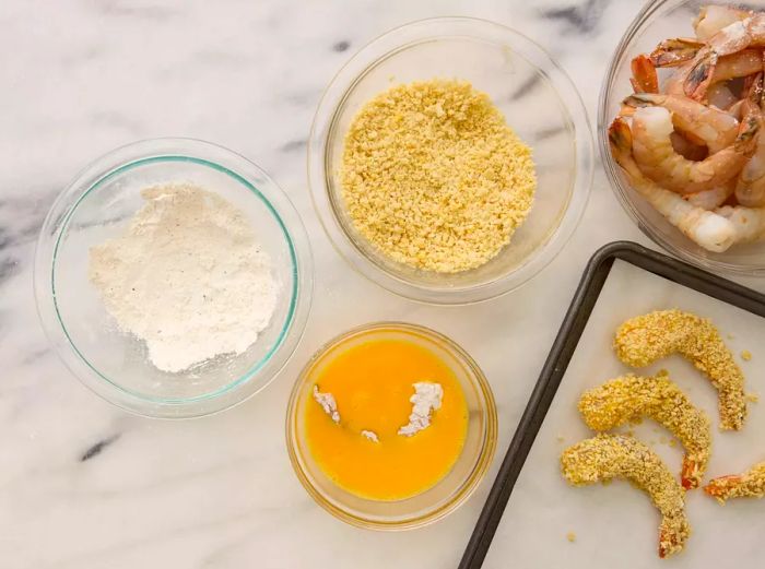 A flour-coated shrimp dipped in a bowl of beaten eggs, next to a small bowl of flour, a bowl of breadcrumbs, and a baking sheet lined with parchment paper, holding the breaded shrimp
