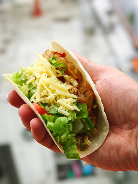 A hand holds a taco packed with lettuce, shredded cheese, grilled chicken, tomatoes, and onions
