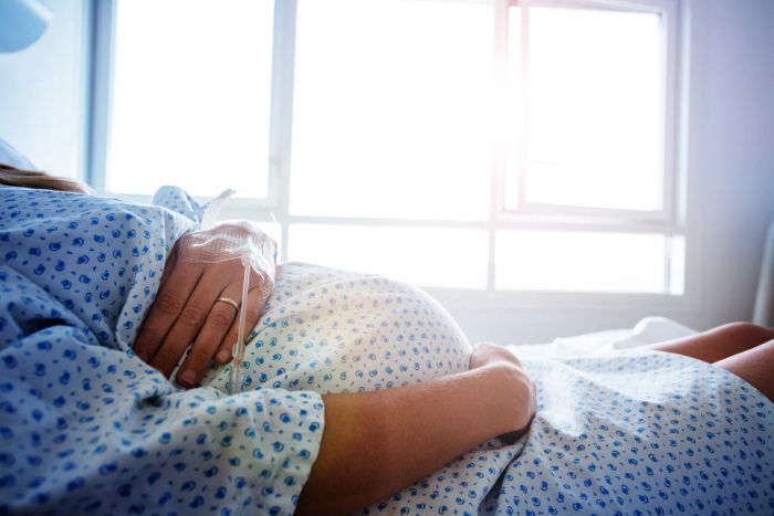 A pregnant person is resting in a hospital bed, tenderly holding their belly, with soft sunlight streaming through a window behind them.