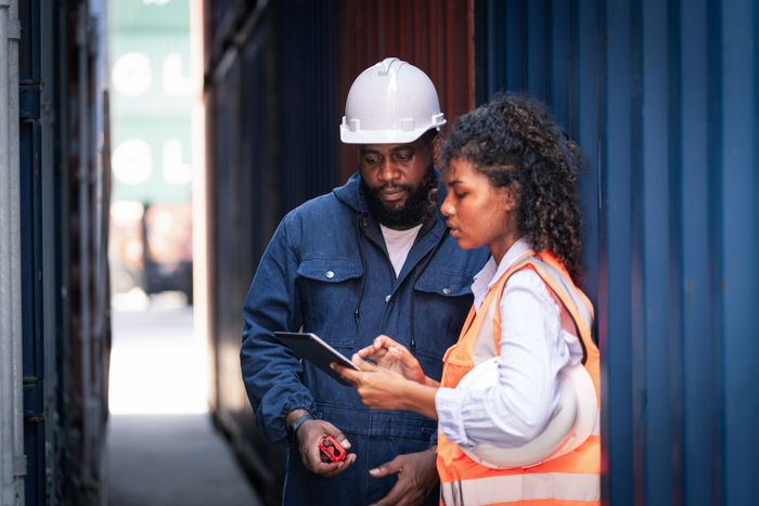 Two employees in a shipping yard are deep in conversation. One is holding a tablet and wearing a safety vest, while the other sports a hard hat and carries a measuring tape.