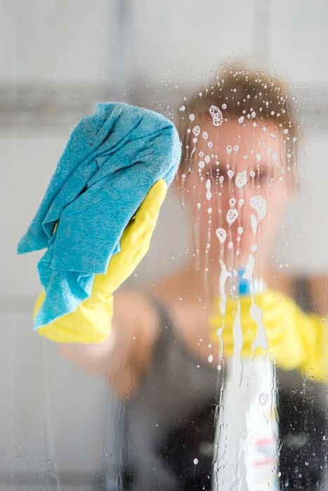A person wearing yellow rubber gloves cleans a glass surface with a blue cloth and a spray bottle