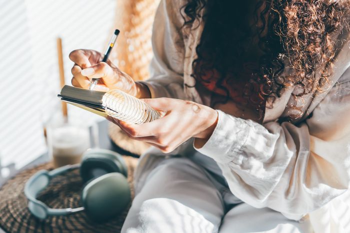 A person with curly hair writes in a journal indoors, with headphones and a candle nearby