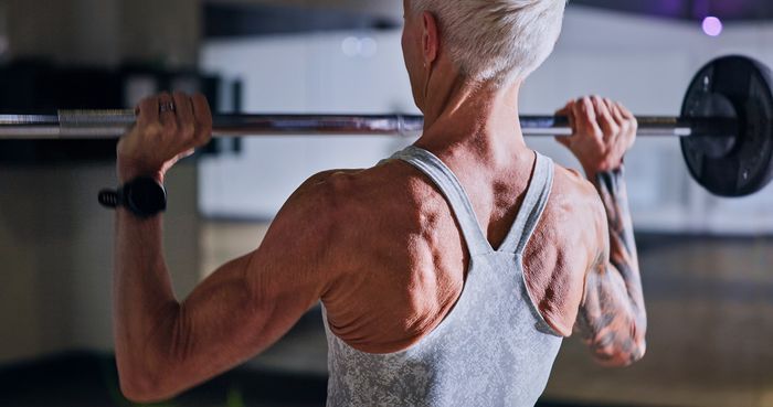A person in a gym lifting weights, wearing a tank top. Their muscular back and focused expression highlight their dedication to strength training.