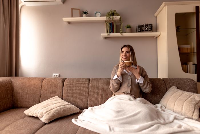 A woman in a cozy sweater holds a cup of coffee while sitting on a couch draped with a blanket. She smiles contently in a warmly decorated living room.