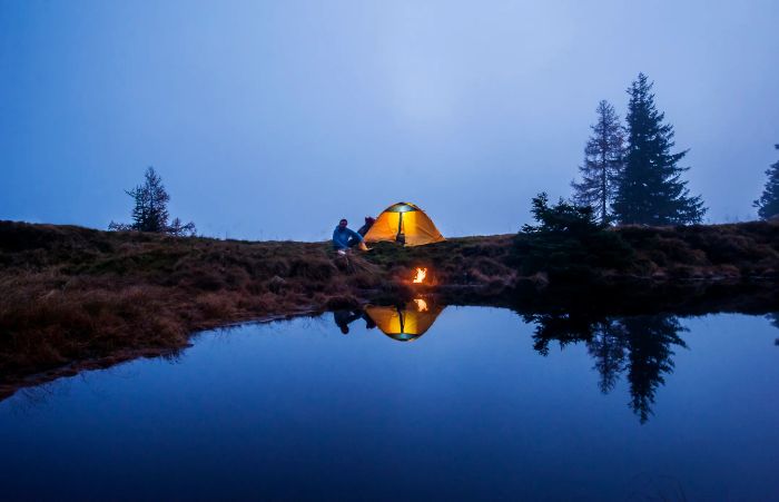 A campsite with a tent and campfire near a serene, reflective pond; a person sits beside the fire under a clear sky, surrounded by towering trees.