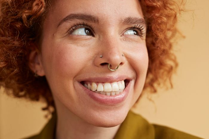 A person with curly hair, a nose ring, and a septum piercing smiles while looking away