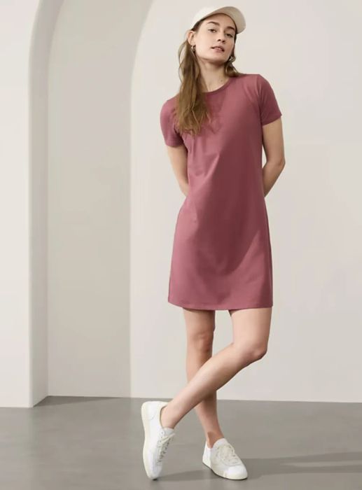 A model wearing a casual short-sleeve dress and white sneakers strikes a pose under an arched doorway, accessorized with a white cap.