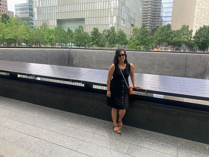 A reviewer poses in front of a memorial structure featuring engraved names, standing on a paved path with modern buildings and trees in the background