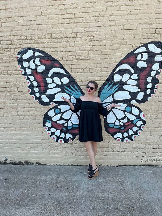 A woman wearing a black off-shoulder dress strikes a pose in front of a butterfly-wing mural on a brick wall