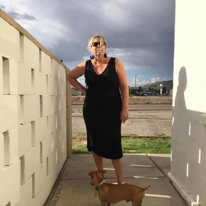 A woman in a black sleeveless dress stands holding a dog on a leash