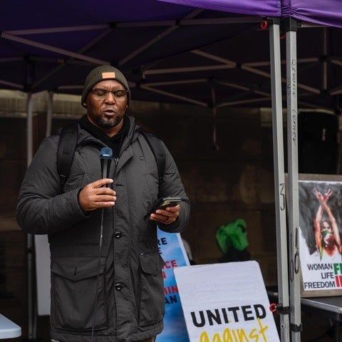 A person in winter attire addresses a protest under a tent, with nearby signs advocating for women’s freedom and unity.