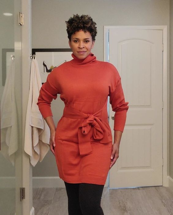 A person wearing a fashionable, belted sweater dress with long sleeves and leggings, standing in a bathroom