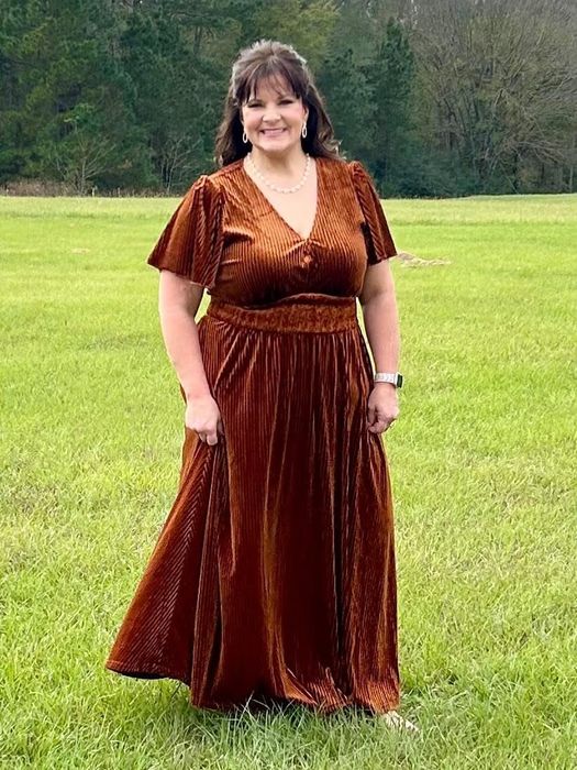 A person outdoors, wearing a flowing brown velvet dress with short sleeves, poses gracefully on grass, exuding elegance perfect for shopping features