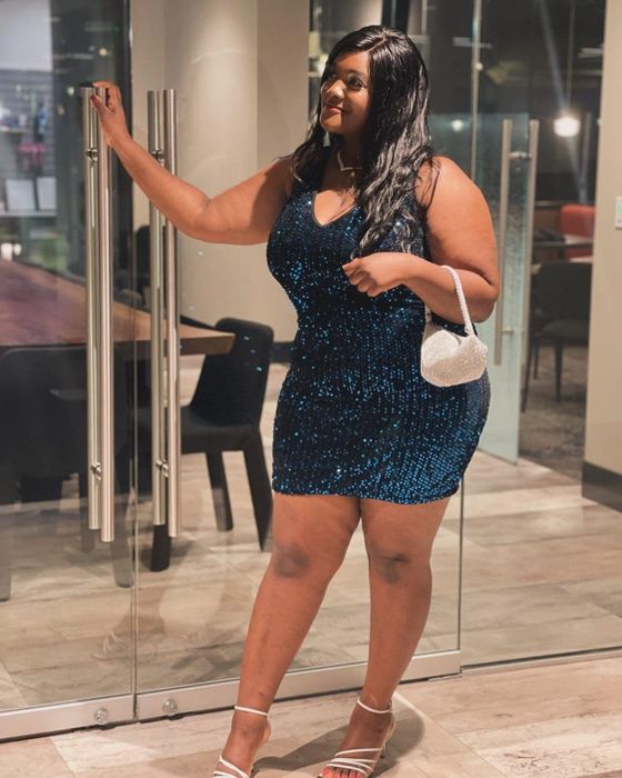 A person confidently poses in a sequined dress, holding a white purse, standing gracefully by a glass door.