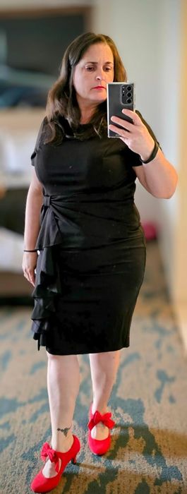 An individual strikes a pose in a chic black dress and vibrant red shoes, capturing a mirror selfie in a well-lit room.