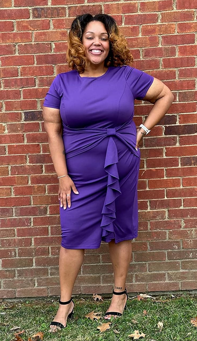 A reviewer smiles brightly in a knee-length, short-sleeved purple dress with ruffled details, paired with black strap heels, standing confidently in front of a rustic brick wall.