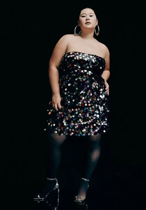 A person confidently poses in a strapless, sequin-embellished dress, accessorized with hoop earrings and metallic heels, against a dark background