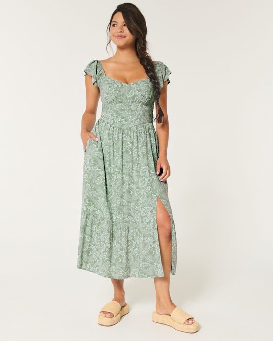 A model wearing a green floral-patterned maxi dress with short ruffled sleeves, a front slit, and tan platform sandals