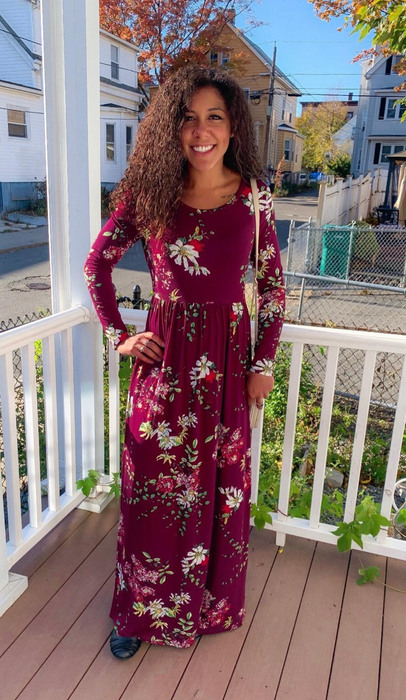 A person smiles on a porch, dressed in a long red floral dress, with houses and trees visible in the background