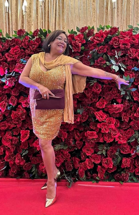 A person radiates joy in a sparkling dress and shawl, posing elegantly against a backdrop of red roses and candles on the red carpet, clutching a stylish clutch.
